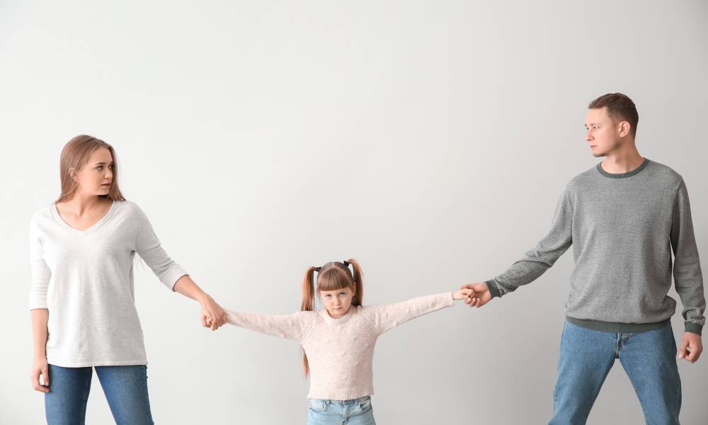 A mom and dad standing on either side of their daughter, both holding one of her hands as they pull her in both directions.