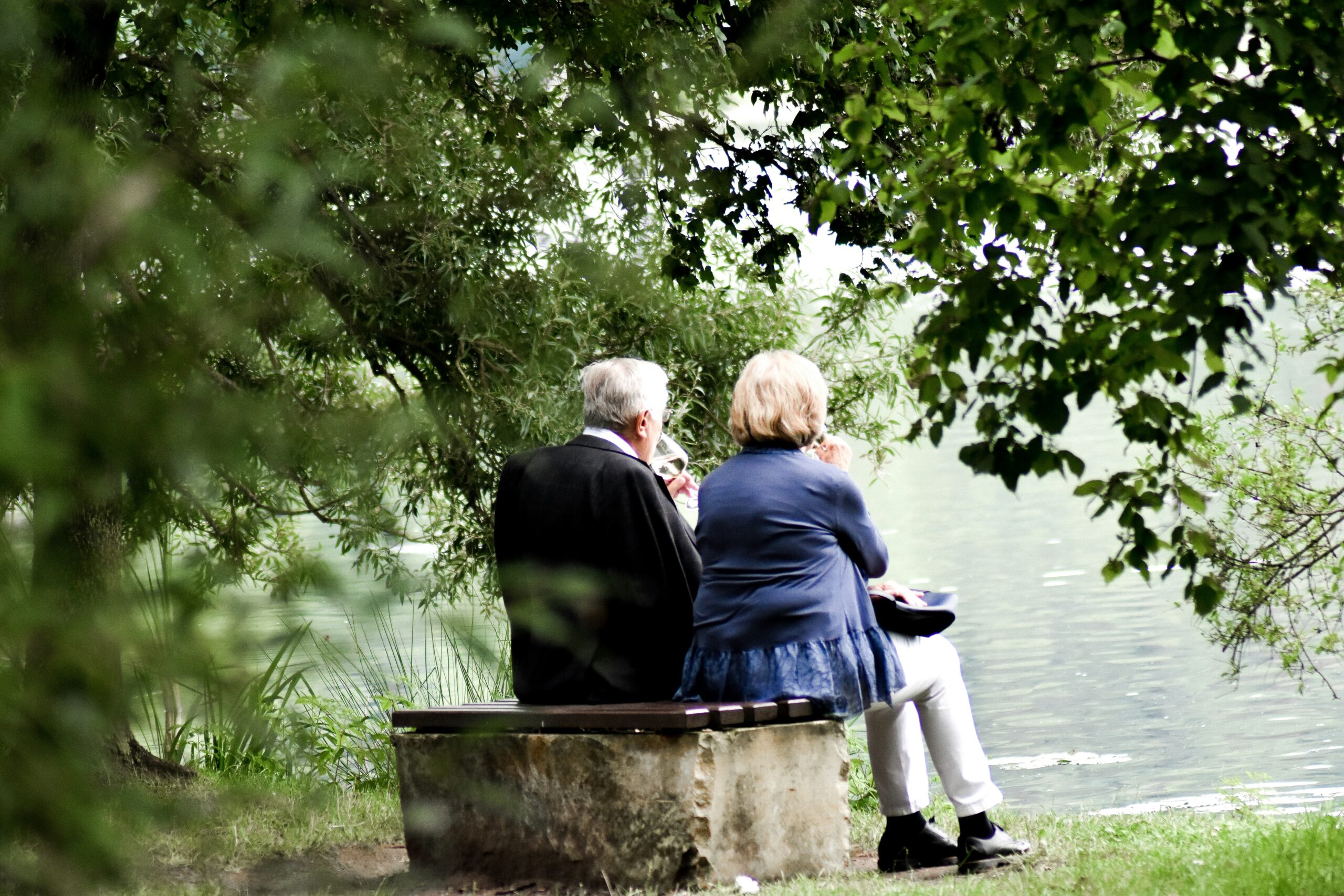 Two people sit beneath leafy trees on a bench.